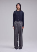 A person stands facing forward in the ISSEY MIYAKE SUEDE LIKE PLEATS, a dark ribbed sweater with a suede-like sheen, paired with wide-leg gray trousers with front flap pockets against a plain light background.