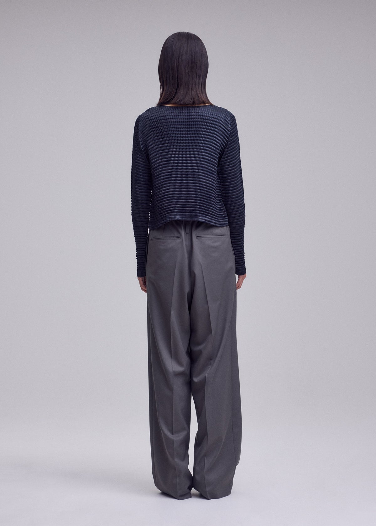 A person with straight dark hair faces away, wearing the ISSEY MIYAKE SUEDE LIKE PLEATS cardigan and loose gray trousers against a plain light gray background. view 6 of 7