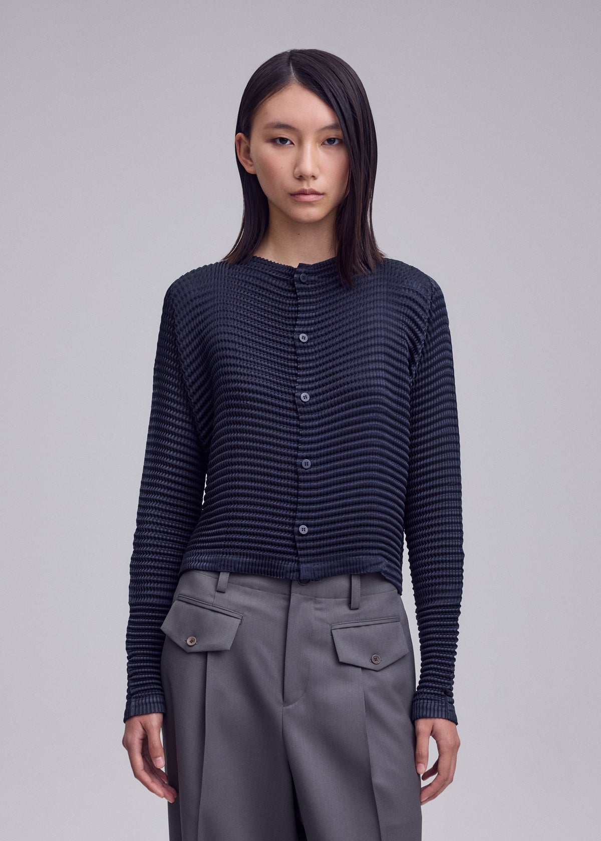 Zoomed view A person with straight dark hair wears the ISSEY MIYAKE SUEDE LIKE PLEATS cardigan and high-waisted gray flap-pocket pants, standing against a plain light background.
