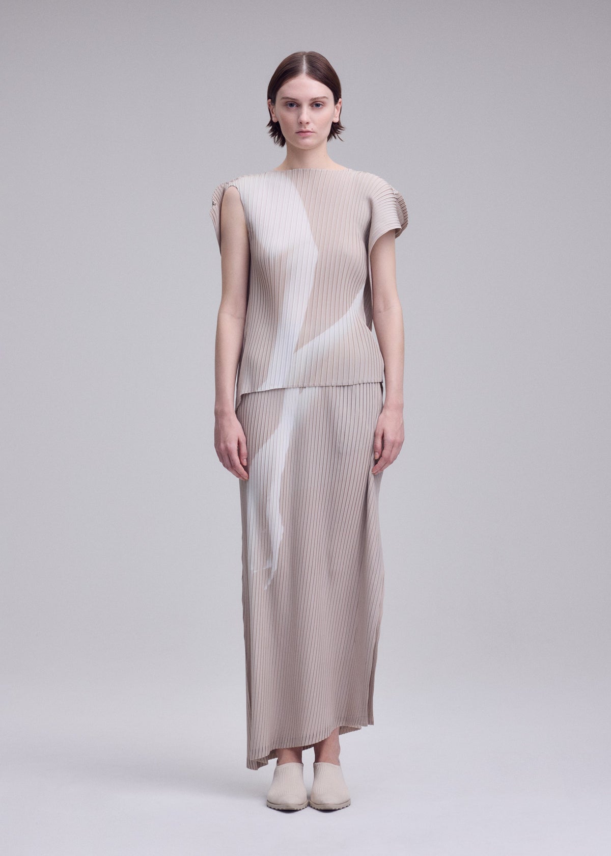 A woman stands facing forward, wearing the ISSEY MIYAKE HANDS OF LOVER PLEATS set—a beige, pleated, short-sleeve top and matching ankle-length skirt featuring abstract white patterns evocative of a monochrome film. Crafted from polyester crepe de chine with inkjet printing, she completes the look with beige shoes against a plain light gray background. view 4 of 7