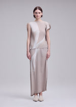 A woman stands facing forward, wearing the ISSEY MIYAKE HANDS OF LOVER PLEATS set—a beige, pleated, short-sleeve top and matching ankle-length skirt featuring abstract white patterns evocative of a monochrome film. Crafted from polyester crepe de chine with inkjet printing, she completes the look with beige shoes against a plain light gray background.