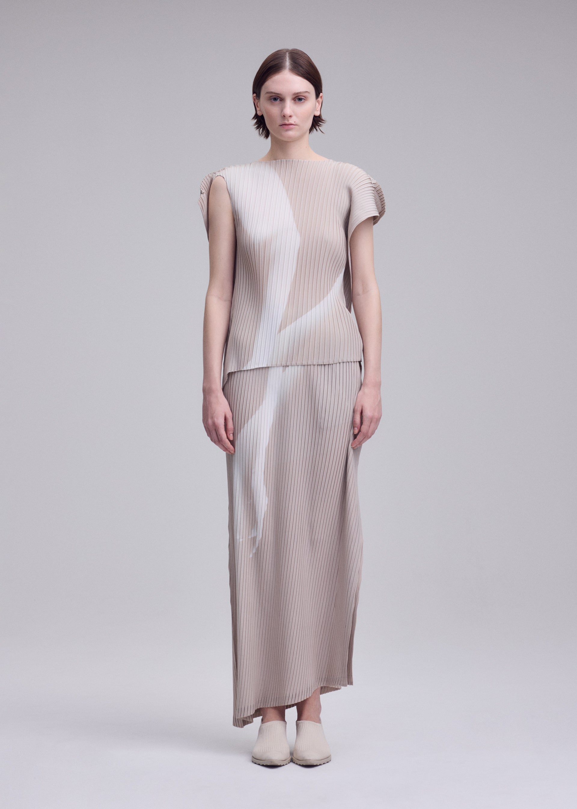 Zoomed view A woman stands facing forward, wearing the ISSEY MIYAKE HANDS OF LOVER PLEATS set—a beige, pleated, short-sleeve top and matching ankle-length skirt featuring abstract white patterns evocative of a monochrome film. Crafted from polyester crepe de chine with inkjet printing, she completes the look with beige shoes against a plain light gray background.