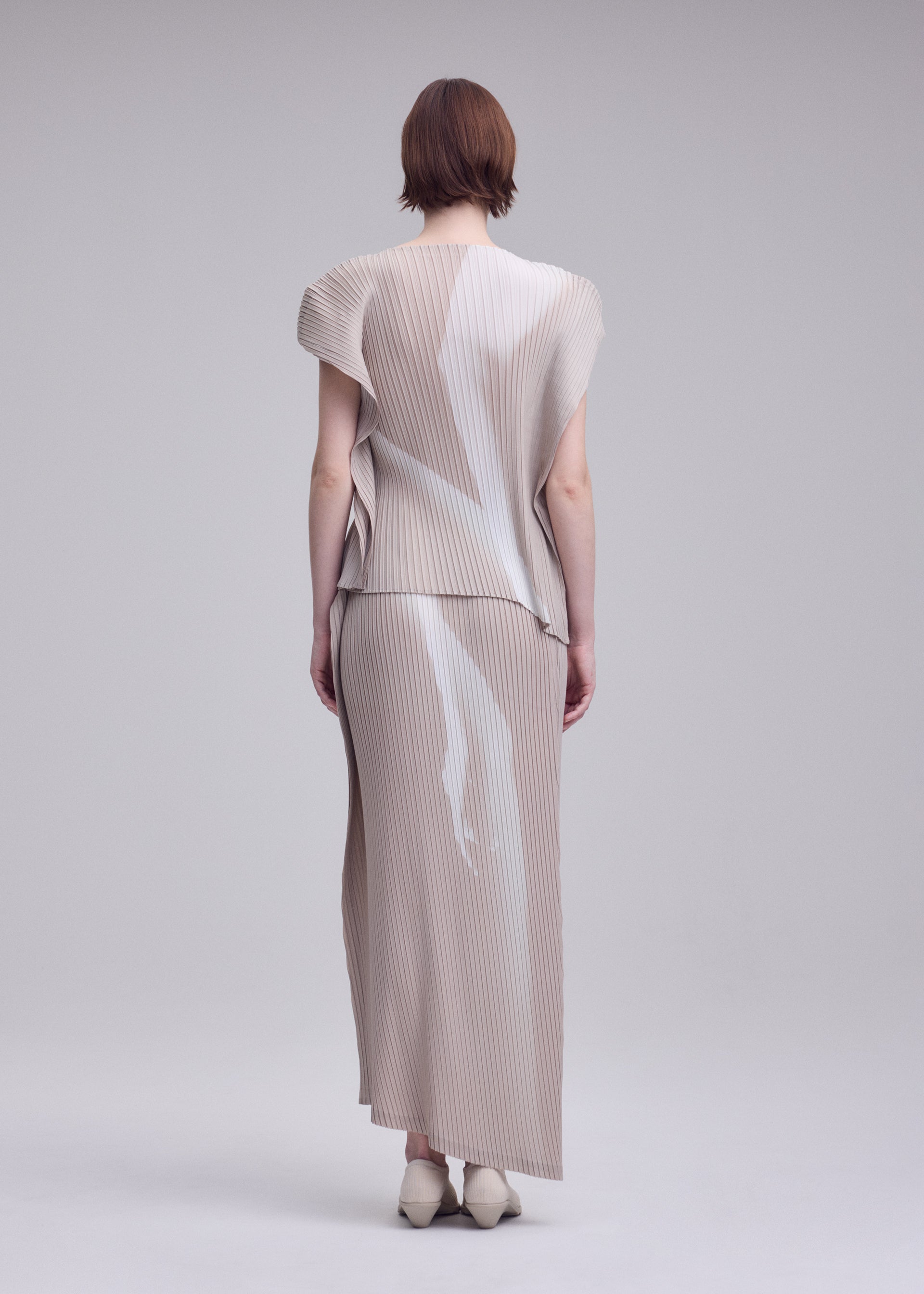 Zoomed view A person with short brown hair, seen from behind, wears the ISSEY MIYAKE HANDS OF LOVER PLEATS set—a beige pleated skirt and top adorned with abstract white patterns evocative of a monochrome film—while standing against a plain light gray background.