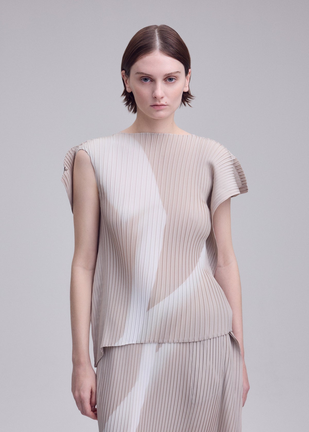 A person with short brown hair wears the ISSEY MIYAKE HANDS OF LOVER PLEATS set— a beige, pleated top and skirt crafted from polyester crepe de chine featuring subtle white patterns inspired by monochrome film motifs—standing against a plain light background. view 6 of 7