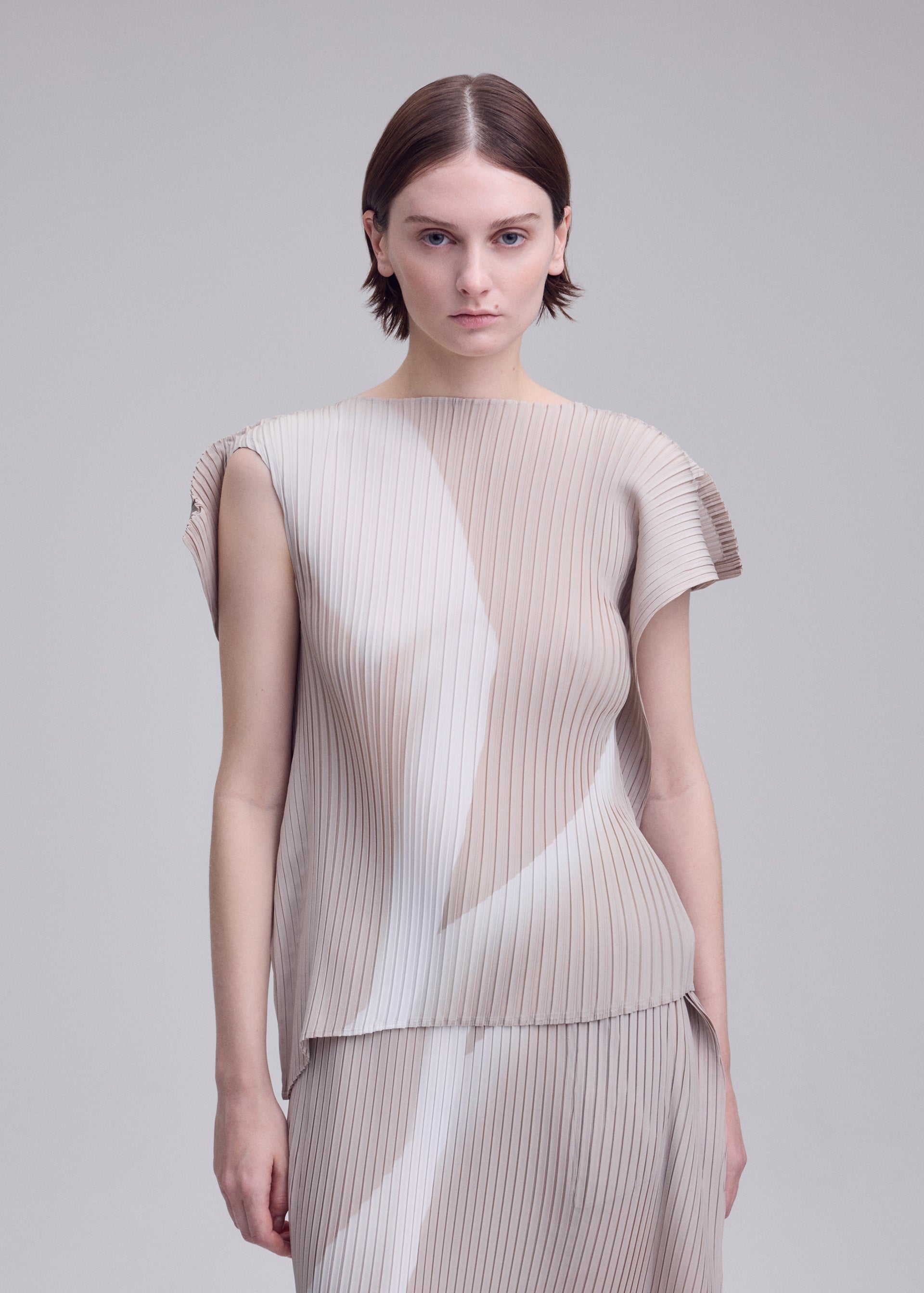 Zoomed view A person with short brown hair wears the ISSEY MIYAKE HANDS OF LOVER PLEATS set— a beige, pleated top and skirt crafted from polyester crepe de chine featuring subtle white patterns inspired by monochrome film motifs—standing against a plain light background.