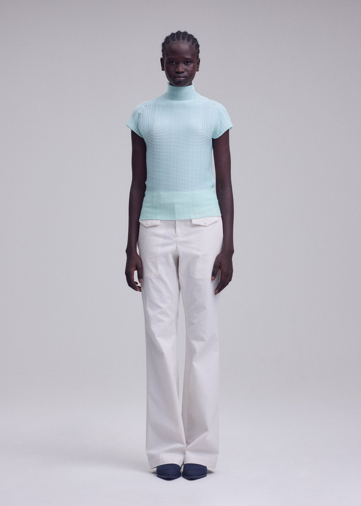 A person with neat braids stands against a plain light background, wearing the ISSEY MIYAKE WOOLY PLEATS-68 high-neck stretchy top with French sleeves in light blue and white recycled polyester wide-leg pants, paired with dark shoes. view 5 of 6