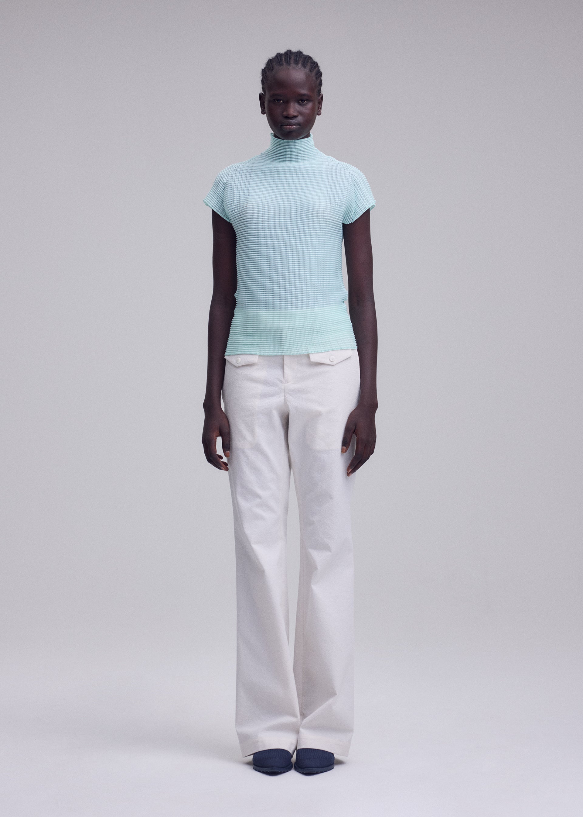 Zoomed view A person with neat braids stands against a plain light background, wearing the ISSEY MIYAKE WOOLY PLEATS-68 high-neck stretchy top with French sleeves in light blue and white recycled polyester wide-leg pants, paired with dark shoes.