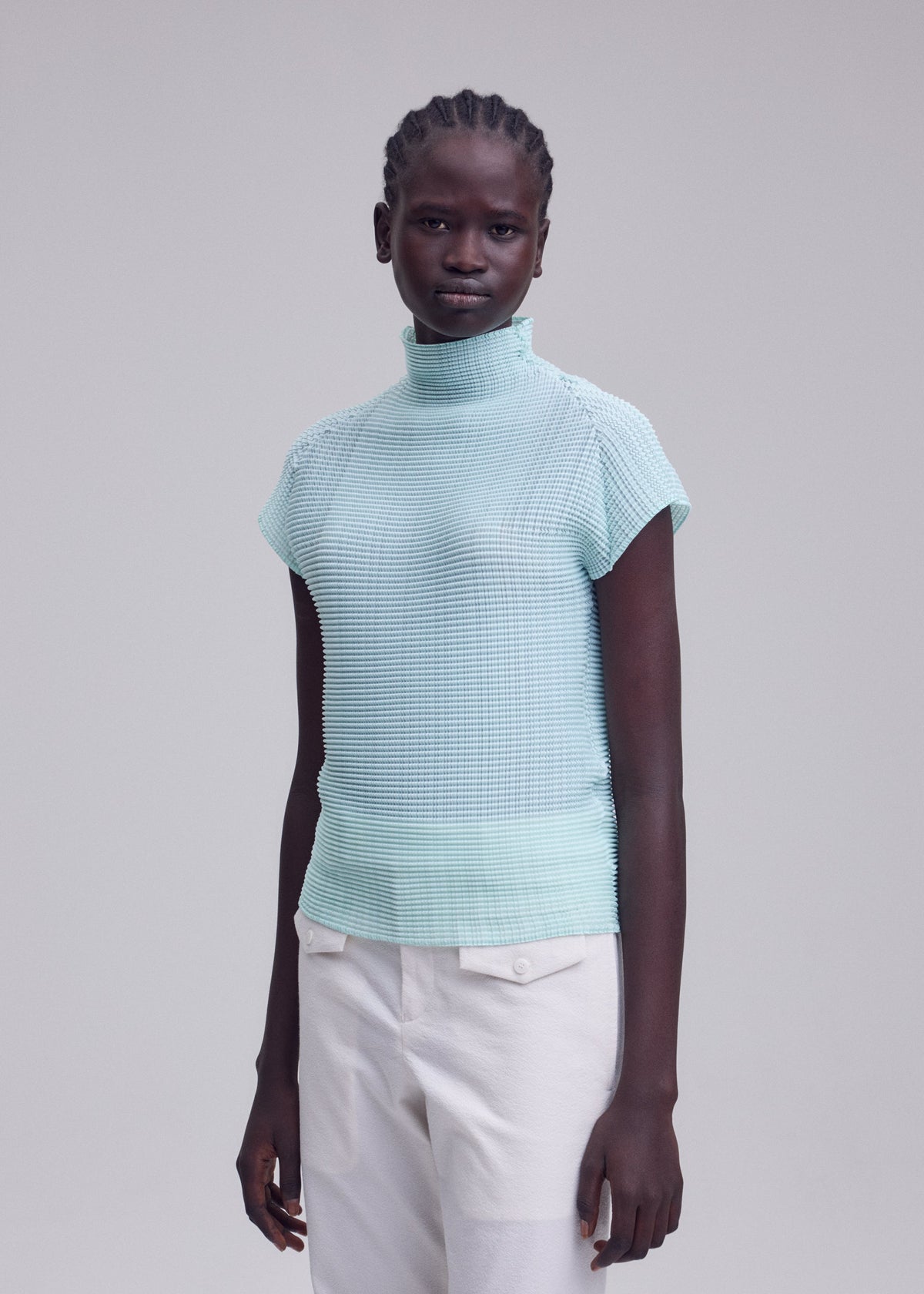 A person with short braided hair models the ISSEY MIYAKE WOOLY PLEATS-68, a light blue stretchy high-neck top with French sleeves, paired with off-white pants against a plain light gray background. view 6 of 6