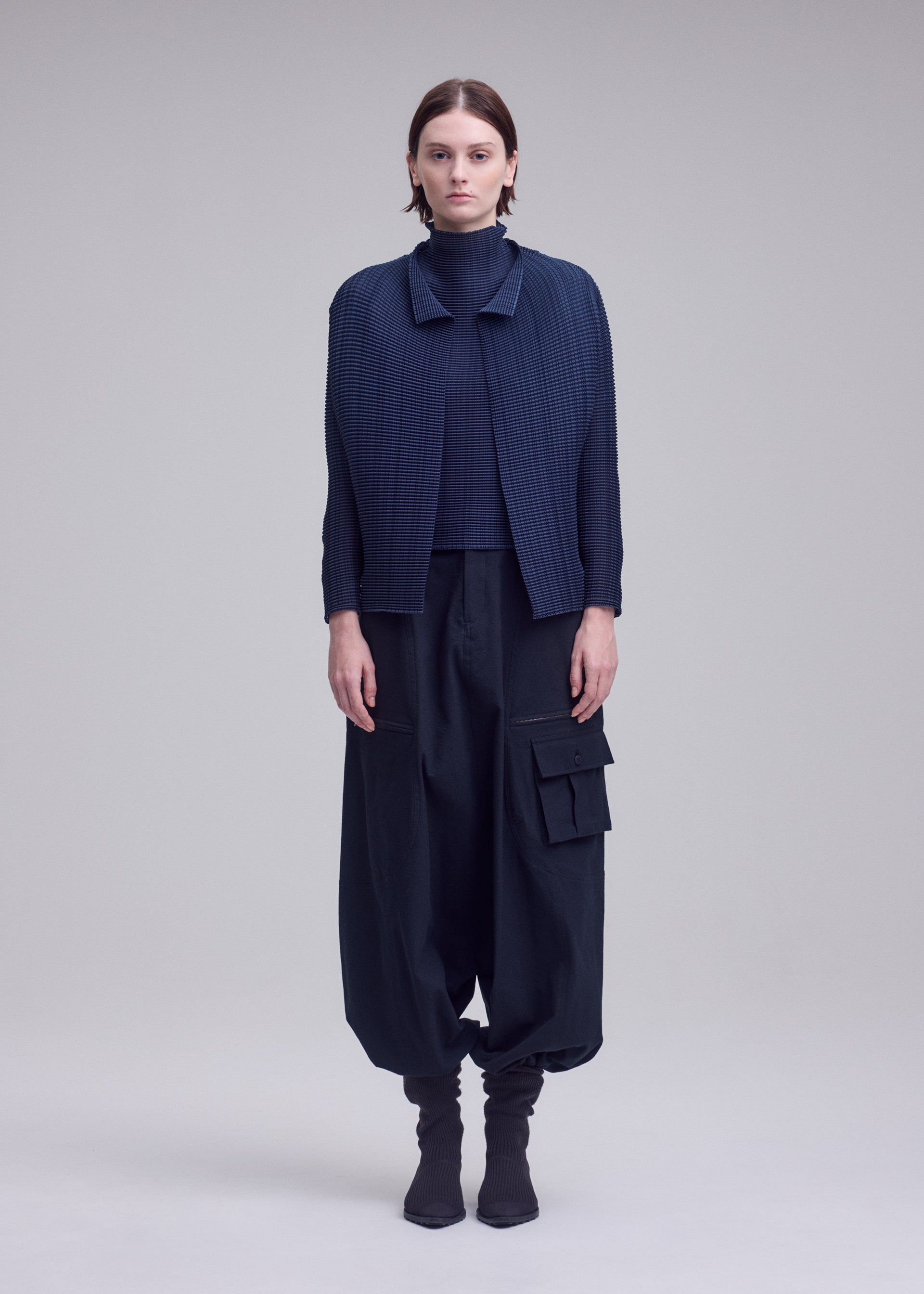 Zoomed view A person faces forward in a plain, light background, wearing ISSEY MIYAKE's WOOLY PLEATS-68 navy checkered pleated cardigan over a turtleneck, with loose dark cargo pants and black socks.