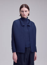 A woman with short brown hair models ISSEY MIYAKE’s WOOLY PLEATS-68: a navy pleated cardigan over a matching top and dark pants, standing against a light background and gazing slightly to the side with a neutral expression.