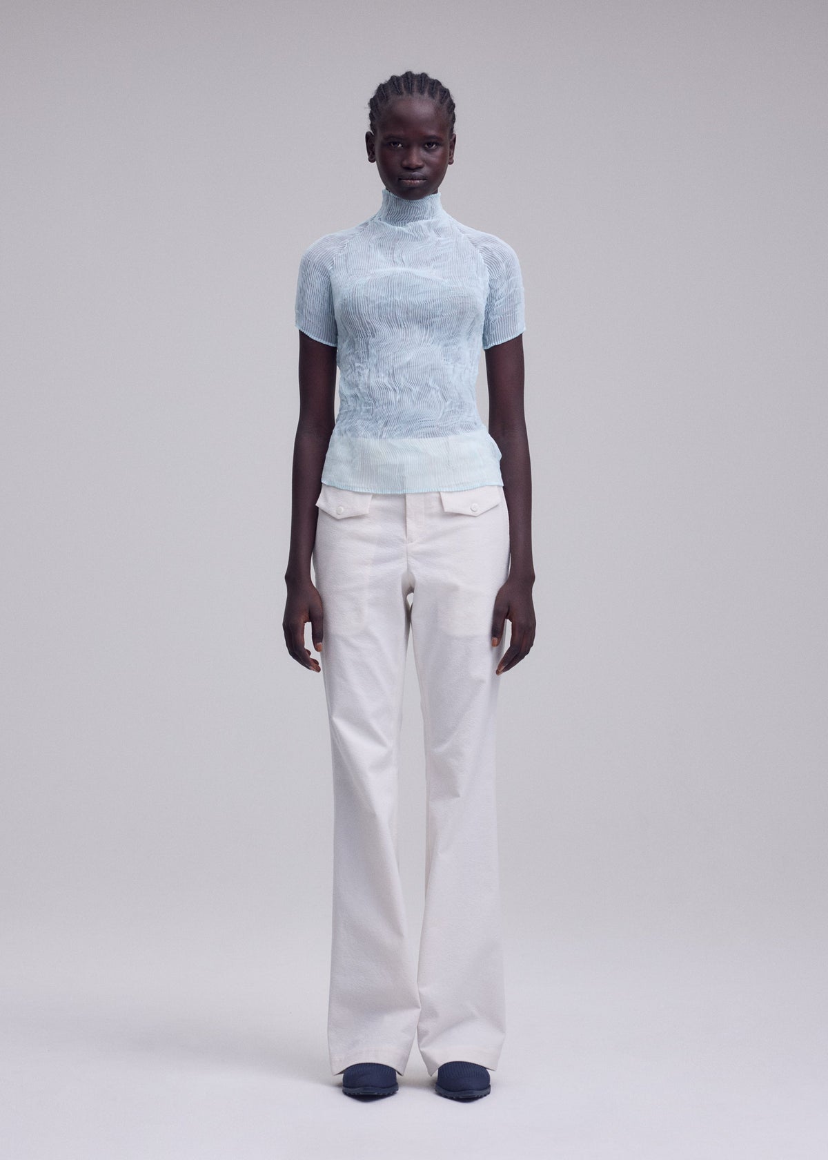 A person stands against a plain light background, facing forward with a neutral expression, wearing the ISSEY MIYAKE CHIFFON TWIST 1 high-neck sheer top in light blue, off-white wide-leg trousers, and black shoes. view 6 of 7
