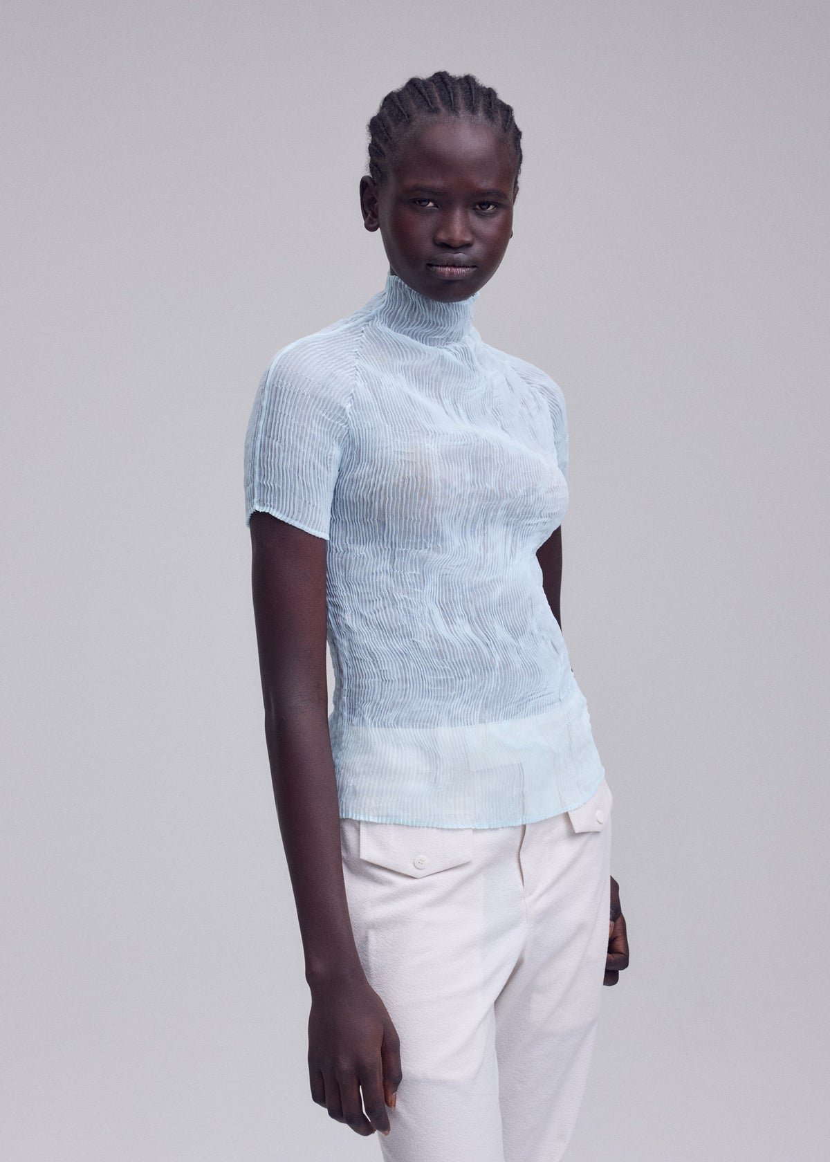 A person with dark skin and braided hair wears the ISSEY MIYAKE CHIFFON TWIST 1, a sheer high-neck light blue top, with light-colored pants, standing against a plain light gray background. view 7 of 7