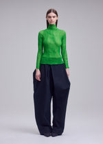 A person with straight, dark hair and a neutral expression stands against a plain background, wearing ISSEY MIYAKE’s CHIFFON TWIST 1: a bright green, high-neck top made from recycled polyester and pleated black wide-leg pants.