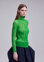 A woman with straight brown hair wears the ISSEY MIYAKE CHIFFON TWIST 1, a sheer bright green long-sleeve high-neck top, and a voluminous black skirt against a plain light gray background.