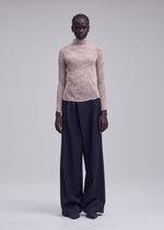A person with neat braids and a neutral expression wears the ISSEY MIYAKE CHIFFON TWIST 2—a sheer beige high-neck top made from recycled polyester—paired with loose, wide-leg black trousers.