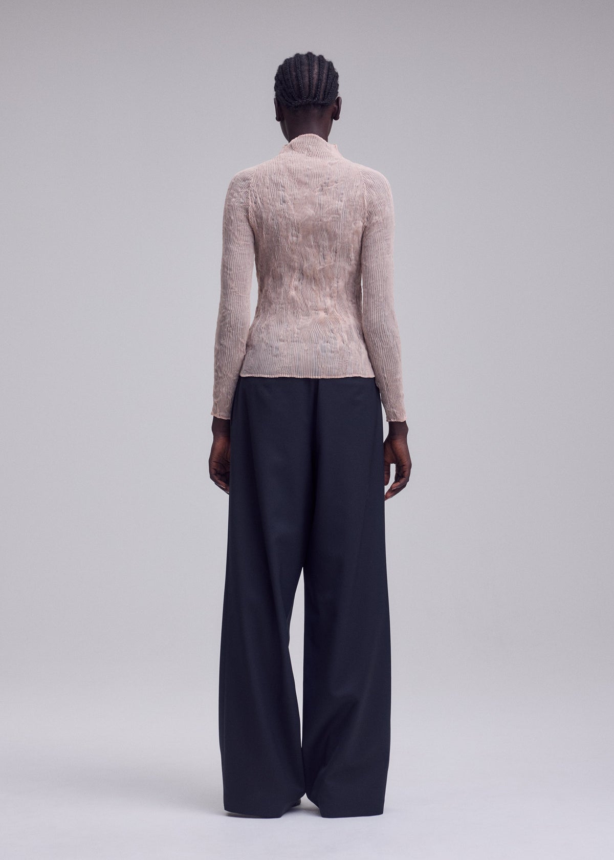 A person with braided hair faces away, wearing the ISSEY MIYAKE CHIFFON TWIST 2 sheer high-neck top and wide-leg navy recycled polyester trousers against a plain light gray background. view 6 of 7