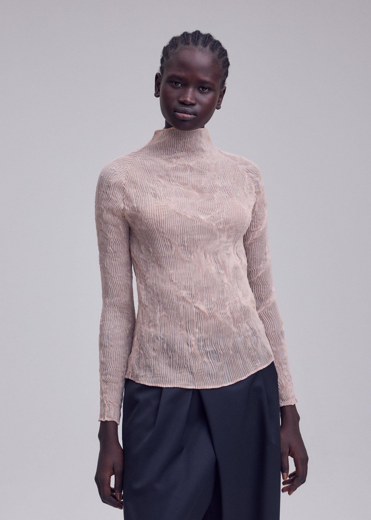 A woman with braided hair wears the ISSEY MIYAKE CHIFFON TWIST 2, a beige, long-sleeve, pleated high-neck top with loose black pants against a plain light gray background. view 7 of 7