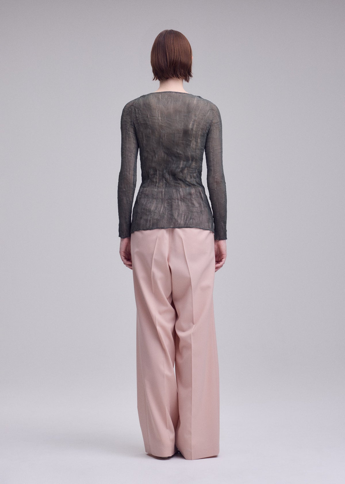 A person faces away, wearing the ISSEY MIYAKE CHIFFON TWIST 2 sheer long-sleeved top with loose, high-waisted blush pink trousers against a plain gray background. view 6 of 7