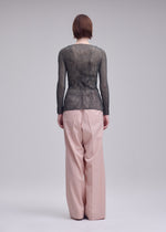 A person faces away, wearing the ISSEY MIYAKE CHIFFON TWIST 2 sheer long-sleeved top with loose, high-waisted blush pink trousers against a plain gray background.