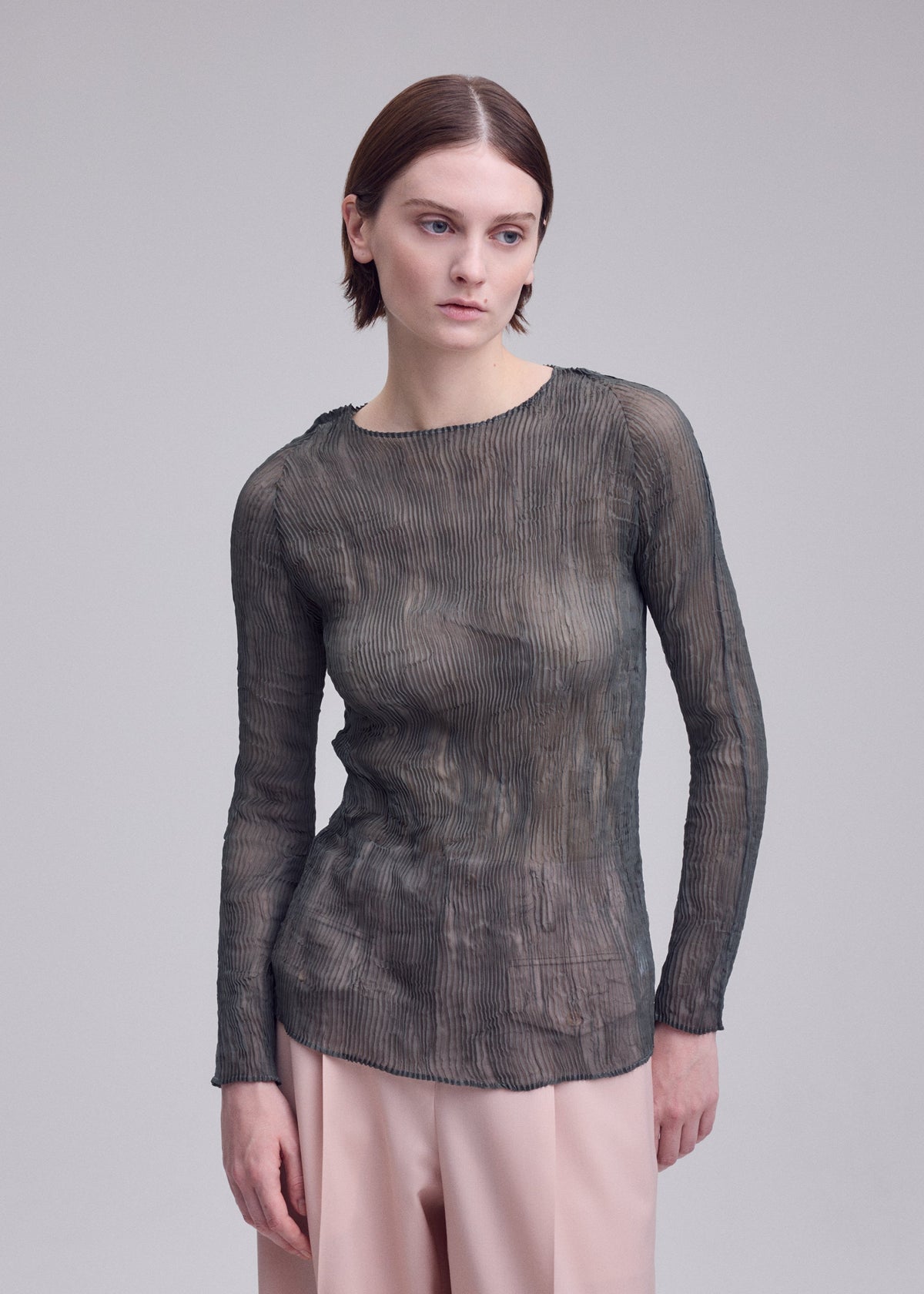 A woman with straight brown hair wears the ISSEY MIYAKE CHIFFON TWIST 2 sheer long-sleeved top and light pink pants, standing against a plain gray background and looking slightly to the side. view 7 of 7