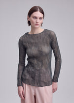 A woman with straight brown hair wears the ISSEY MIYAKE CHIFFON TWIST 2 sheer long-sleeved top and light pink pants, standing against a plain gray background and looking slightly to the side.
