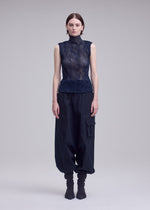 A person stands facing forward, wearing ISSEY MIYAKE’s CHIFFON TWIST 1 high-neck sleeveless top in recycled polyester, loose black cargo pants, and dark boots. The semi-sheer texture contrasts with the plain light gray background.