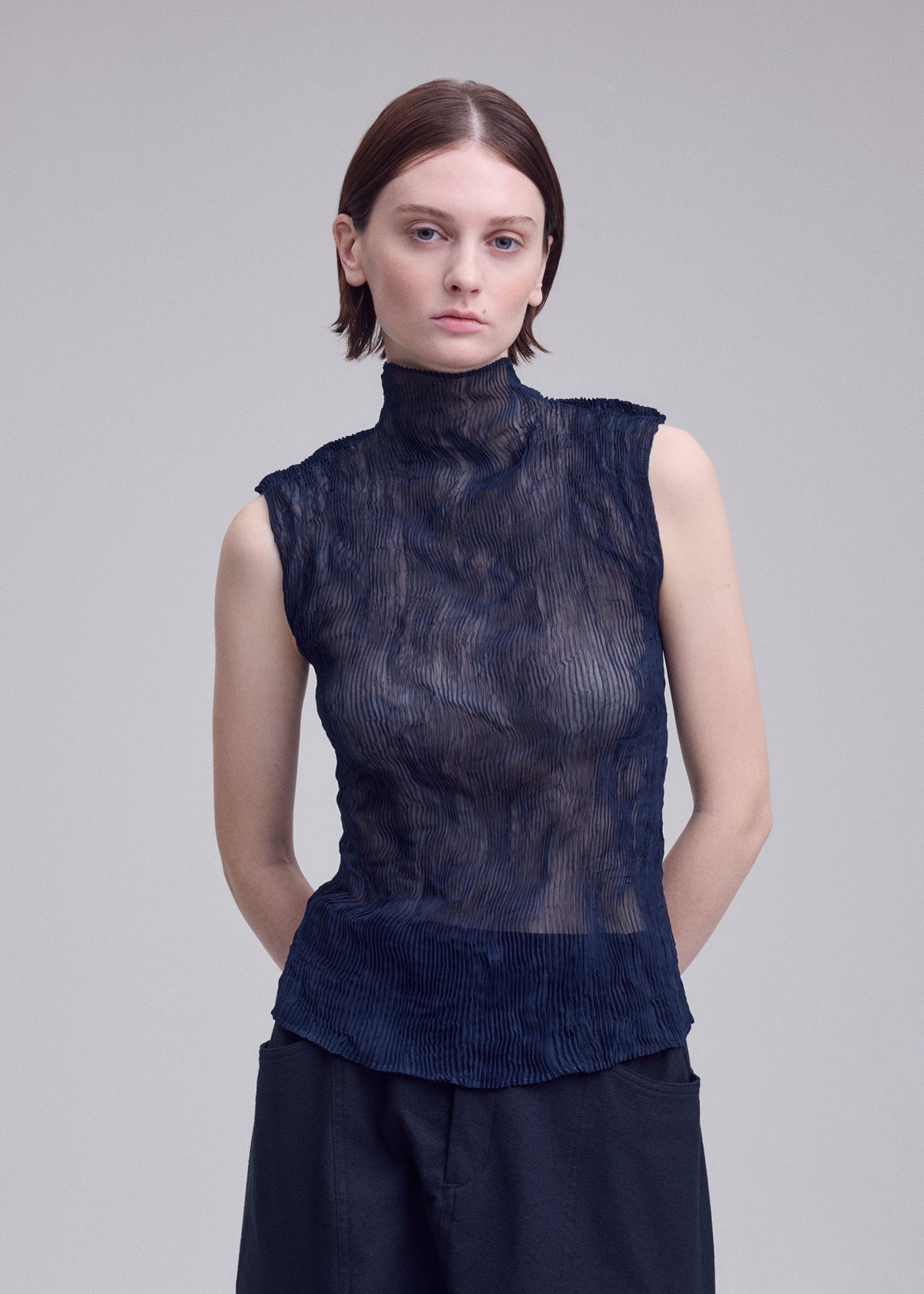 A woman with short brown hair wears the ISSEY MIYAKE CHIFFON TWIST 1, a high-neck sleeveless sheer navy top paired with dark trousers. She stands against a plain light gray background, hands behind her back, looking at the camera. view 7 of 7