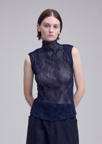 A woman with short brown hair wears the ISSEY MIYAKE CHIFFON TWIST 1, a high-neck sleeveless sheer navy top paired with dark trousers. She stands against a plain light gray background, hands behind her back, looking at the camera.