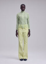 A person stands against a plain background wearing the ISSEY MIYAKE CHIFFON TWIST 1, a sheer light green zip-up cardigan made from recycled polyester, paired with loose pale yellow pleated pants and black shoes.