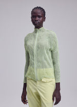 A person with dark skin and braided hair wears ISSEY MIYAKE's CHIFFON TWIST 1, a light green, textured zip-up cardigan made from recycled polyester, paired with pale yellow pleated pants against a plain light gray background.