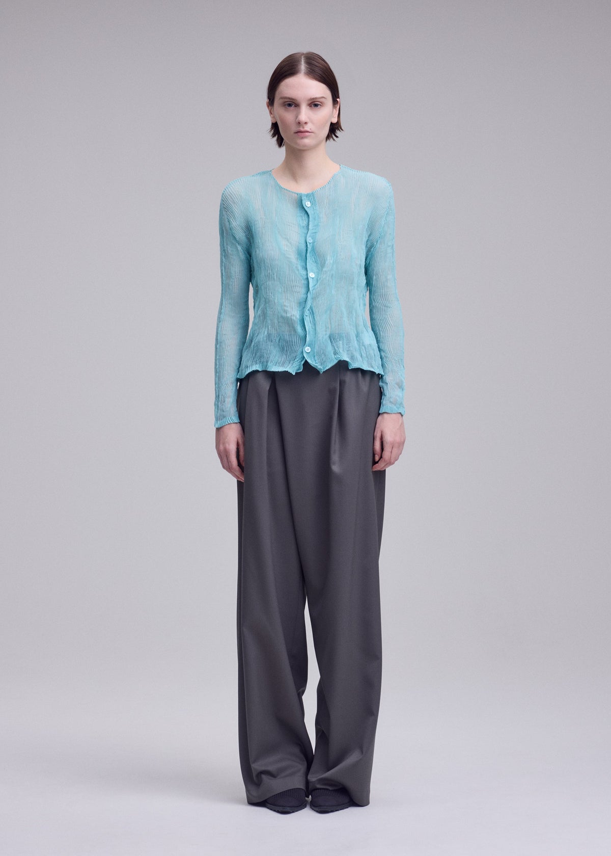 Zoomed view A person stands facing forward in the ISSEY MIYAKE CHIFFON TWIST 2, a light blue semi-sheer cardigan paired with loose gray trousers against a plain, light background.