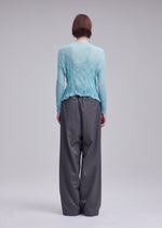 A person with short brown hair stands against a neutral background, wearing the ISSEY MIYAKE CHIFFON TWIST 2 cardigan in light blue and loose-fitting gray trousers.