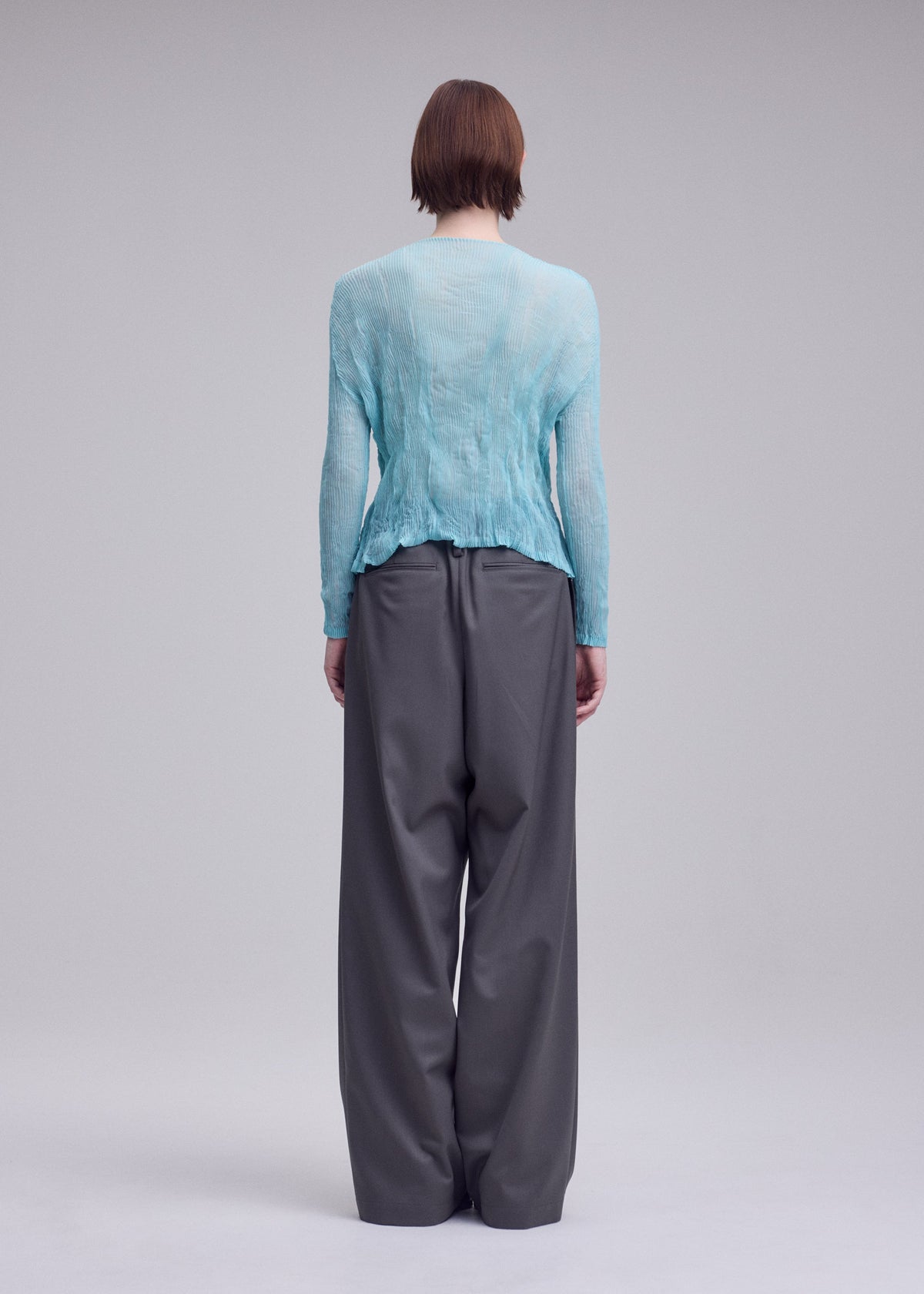 Zoomed view A person with short brown hair stands against a neutral background, wearing the ISSEY MIYAKE CHIFFON TWIST 2 cardigan in light blue and loose-fitting gray trousers.
