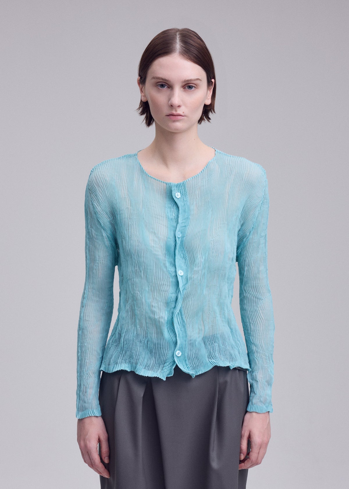 A woman with short brown hair models the CHIFFON TWIST 2 by ISSEY MIYAKE—a sheer light blue buttoned cardigan—paired with loose gray pants, standing against a plain gray background. view 7 of 7