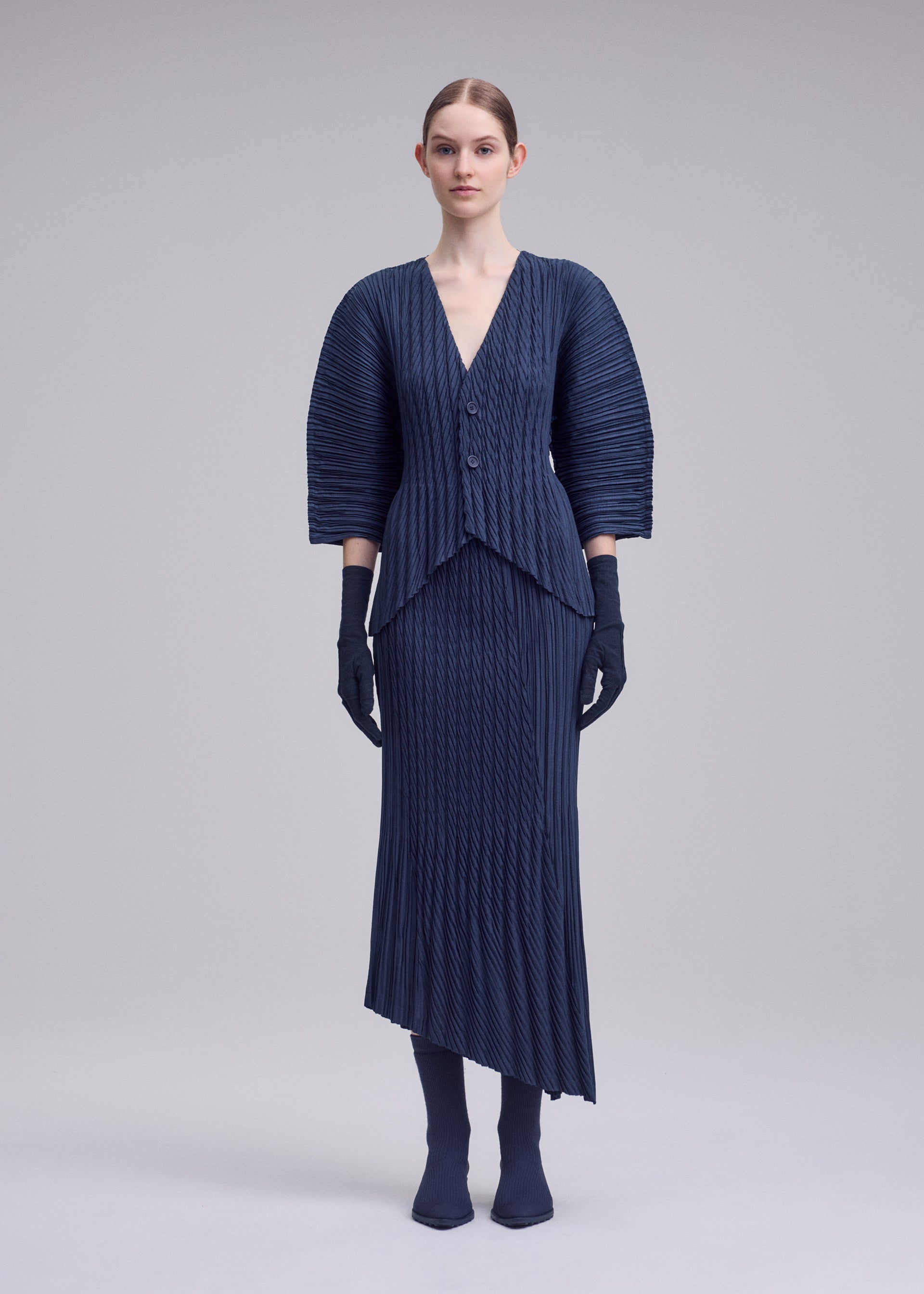 Zoomed view A woman poses against a plain background in the UNFURLING PLEATS set by ISSEY MIYAKE, featuring a pleated navy jacket and skirt with diagonal pleats, structured shoulders, matching gloves, tights, and shoes for a modern textured look.