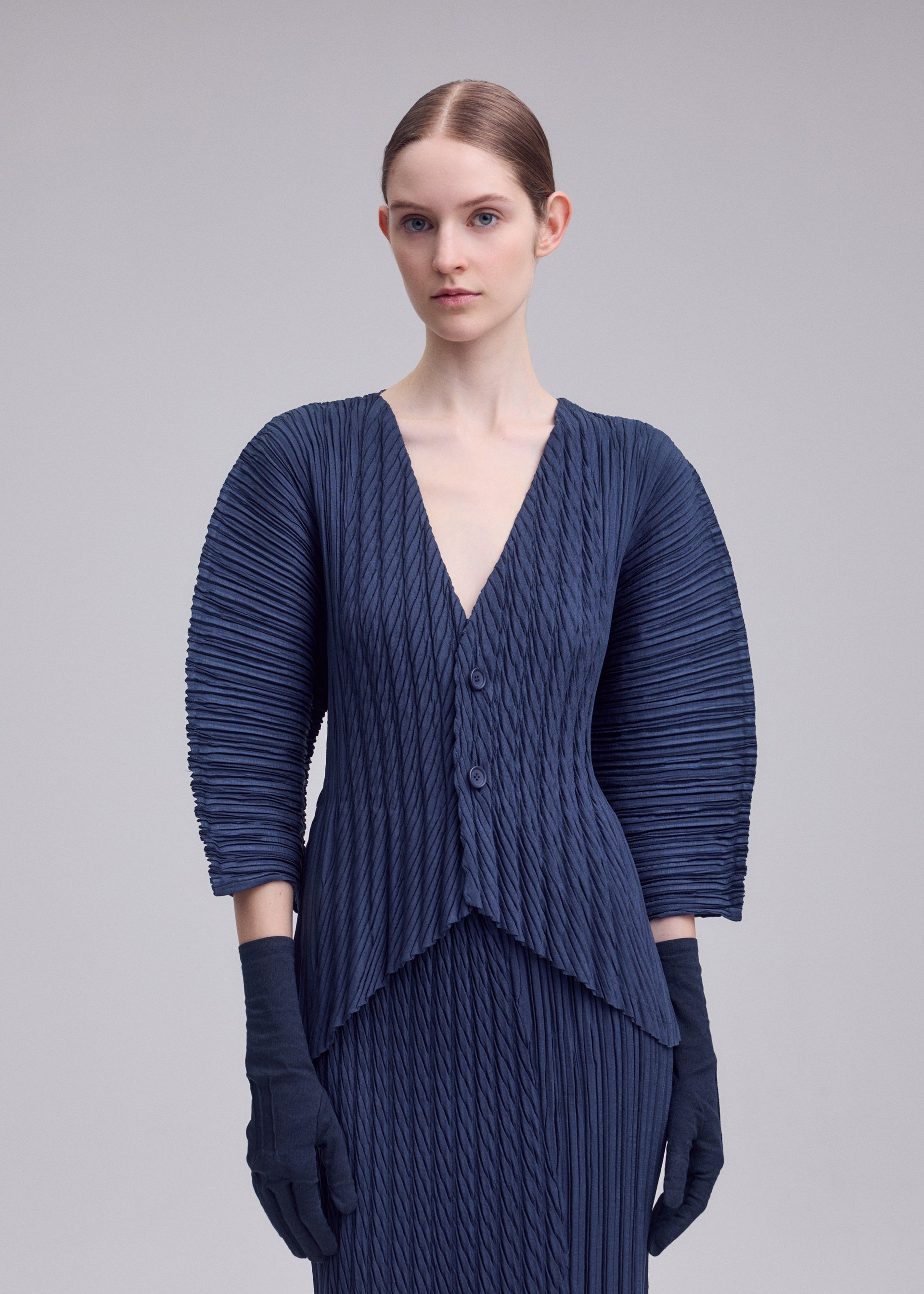 Zoomed view A woman with light skin and straight brown hair pulled back models the ISSEY MIYAKE UNFURLING PLEATS navy V-neck jacket with structured sleeves and matching gloves, standing against a plain light gray background.