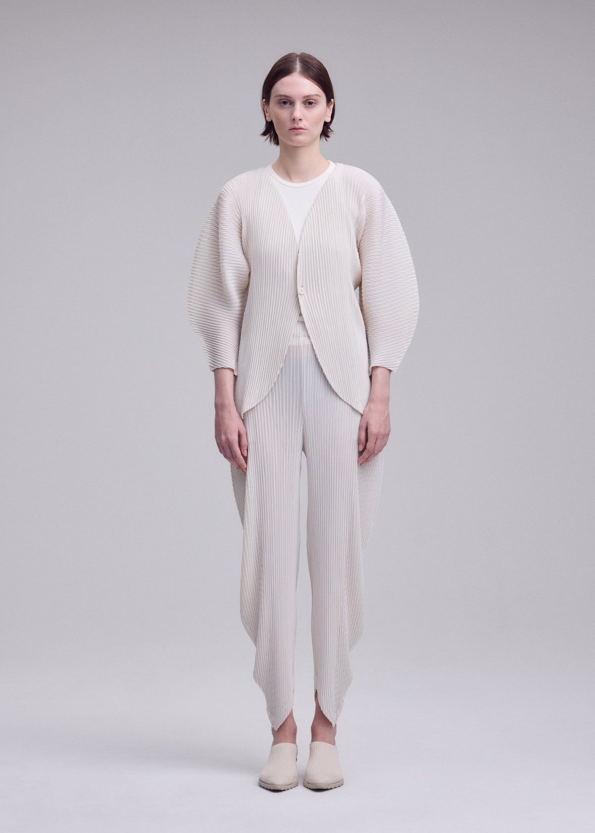 A person stands facing forward, wearing the ISSEY MIYAKE LEAF PLEATS outfit—an off-white textured set featuring pleated pants and a coordinating pleated cardigan—against a plain light gray background. view 5 of 7