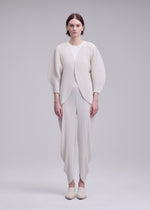 A person stands facing forward, wearing the ISSEY MIYAKE LEAF PLEATS outfit—an off-white textured set featuring pleated pants and a coordinating pleated cardigan—against a plain light gray background.