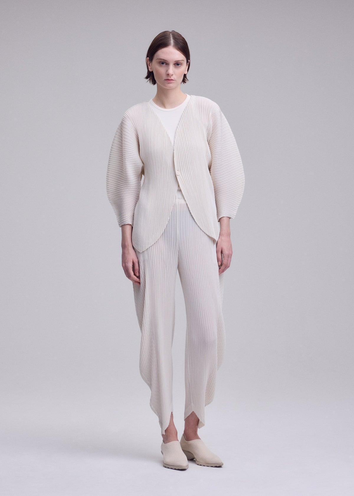 Against a plain background, a person wears the ISSEY MIYAKE LEAF PLEATS set—an off-white, pleated ensemble featuring a double-pleating cardigan with rounded sleeves and coordinating pants. Slip-on shoes complete the minimalist, modern look. view 6 of 7