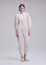 Against a plain background, a person wears the ISSEY MIYAKE LEAF PLEATS set—an off-white, pleated ensemble featuring a double-pleating cardigan with rounded sleeves and coordinating pants. Slip-on shoes complete the minimalist, modern look.