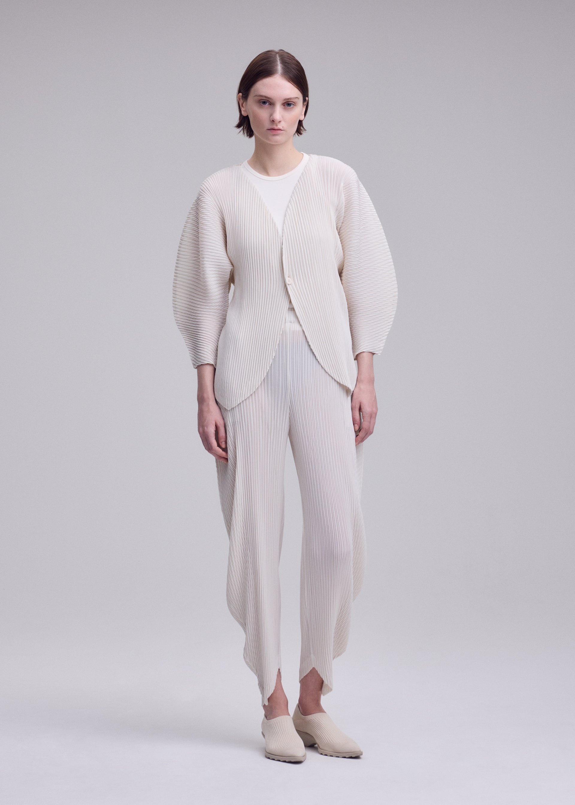 Zoomed view Against a plain background, a person wears the ISSEY MIYAKE LEAF PLEATS set—an off-white, pleated ensemble featuring a double-pleating cardigan with rounded sleeves and coordinating pants. Slip-on shoes complete the minimalist, modern look.