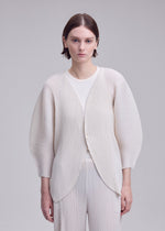 A person with short brown hair stands against a plain light gray background, looking directly at the camera with a neutral expression, wearing the ISSEY MIYAKE LEAF PLEATS textured cream-colored pleated cardigan.