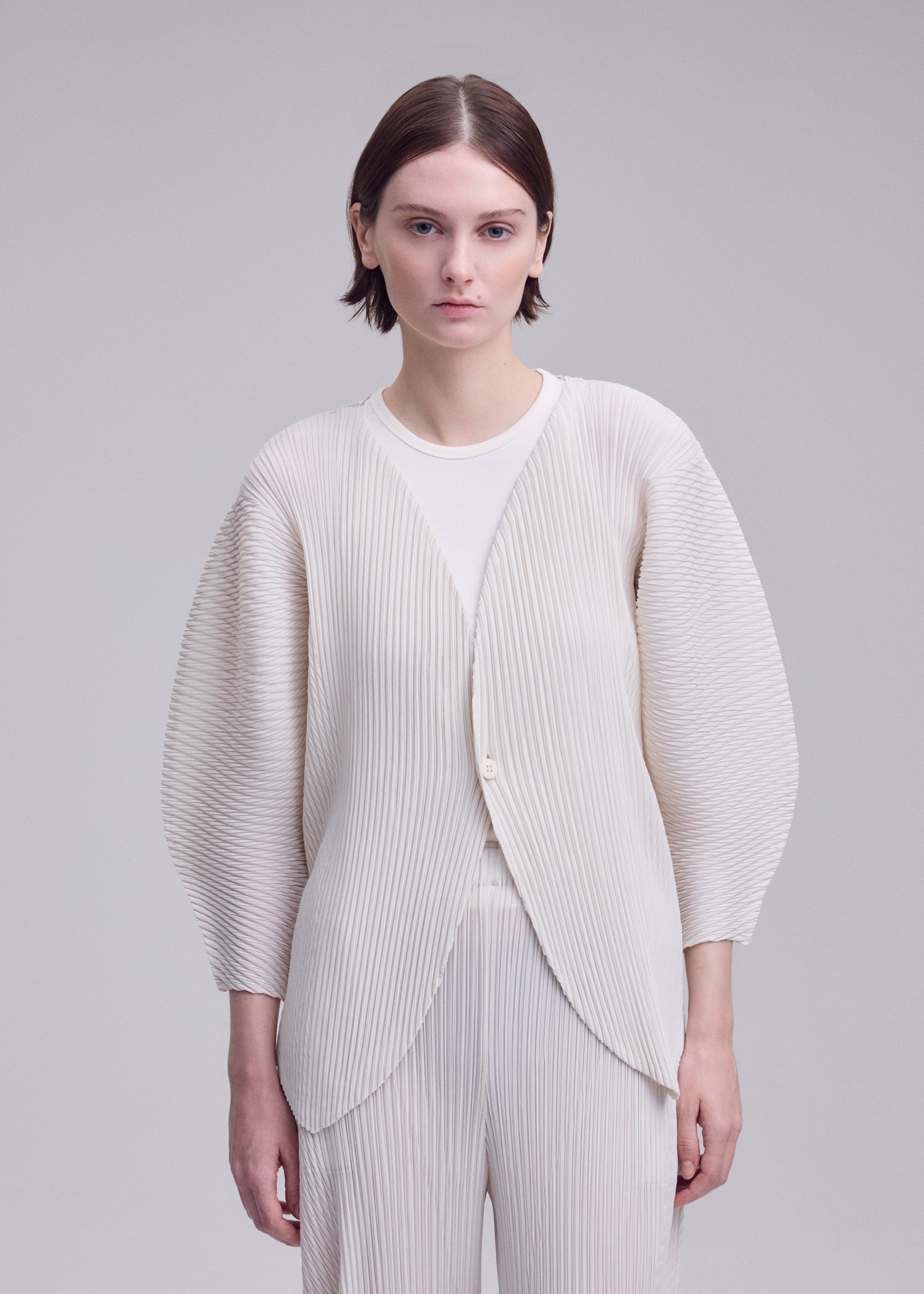 Zoomed view A person with short brown hair stands against a plain light gray background, looking directly at the camera with a neutral expression, wearing the ISSEY MIYAKE LEAF PLEATS textured cream-colored pleated cardigan.