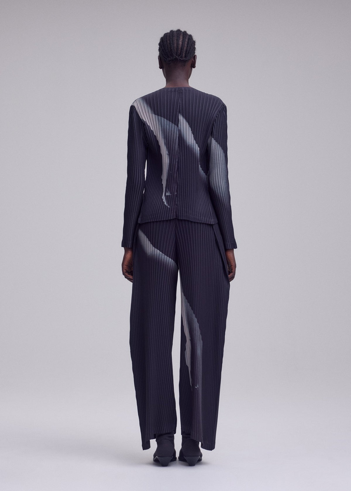 A person stands with their back to the camera, dressed in ISSEY MIYAKE’s HANDS OF LOVER PLEATS suit. The ensemble features a distinctive pinstriped pattern of abstract white streaks on dark polyester crepe de chine. Matching pants and pointed shoes complete the look against a simple, neutral backdrop. view 5 of 7