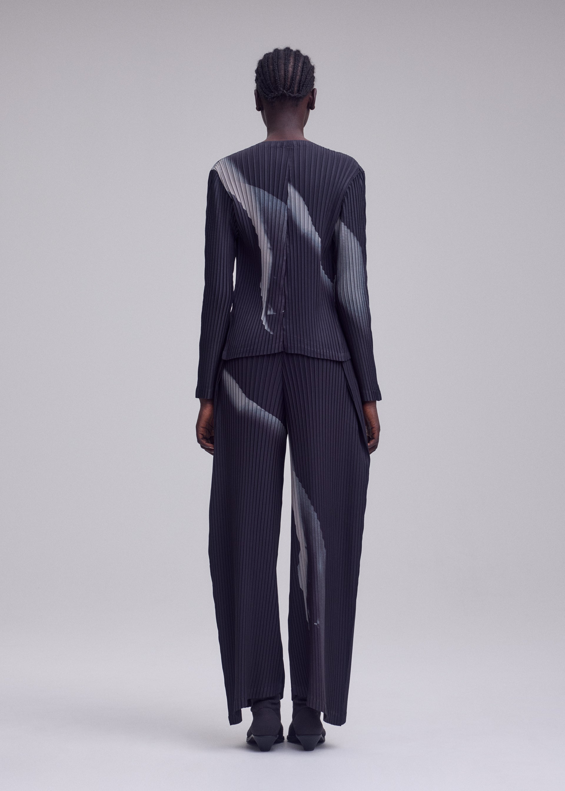 Zoomed view A person stands with their back to the camera, dressed in ISSEY MIYAKE’s HANDS OF LOVER PLEATS suit. The ensemble features a distinctive pinstriped pattern of abstract white streaks on dark polyester crepe de chine. Matching pants and pointed shoes complete the look against a simple, neutral backdrop.
