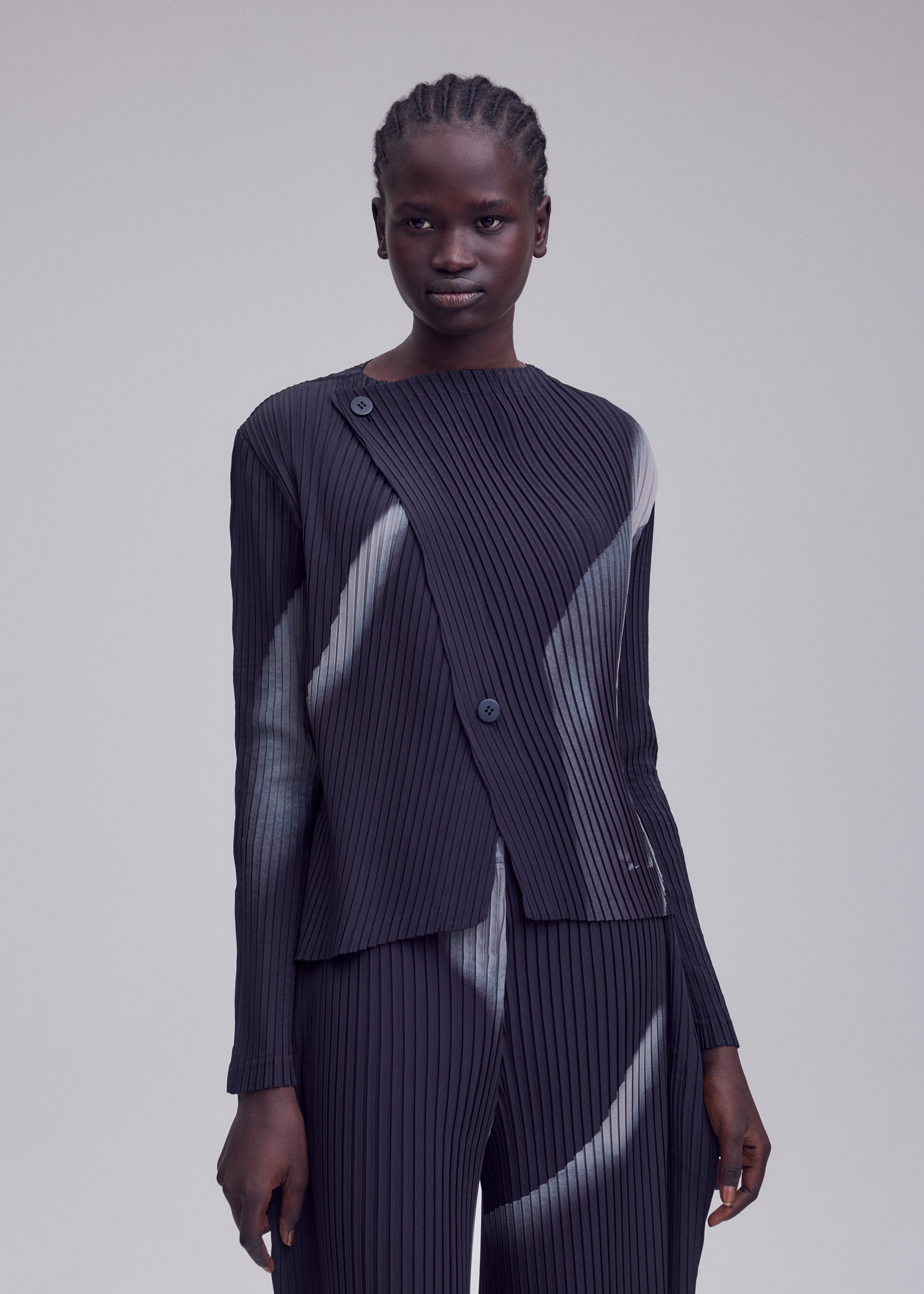 Zoomed view A person with braided hair models the HANDS OF LOVER PLEATS set by ISSEY MIYAKE, featuring a pleated, long-sleeve top and matching pants made from dark polyester crepe de chine adorned with light abstract streaks in an original inkjet-printed pattern, standing against a plain light gray background.