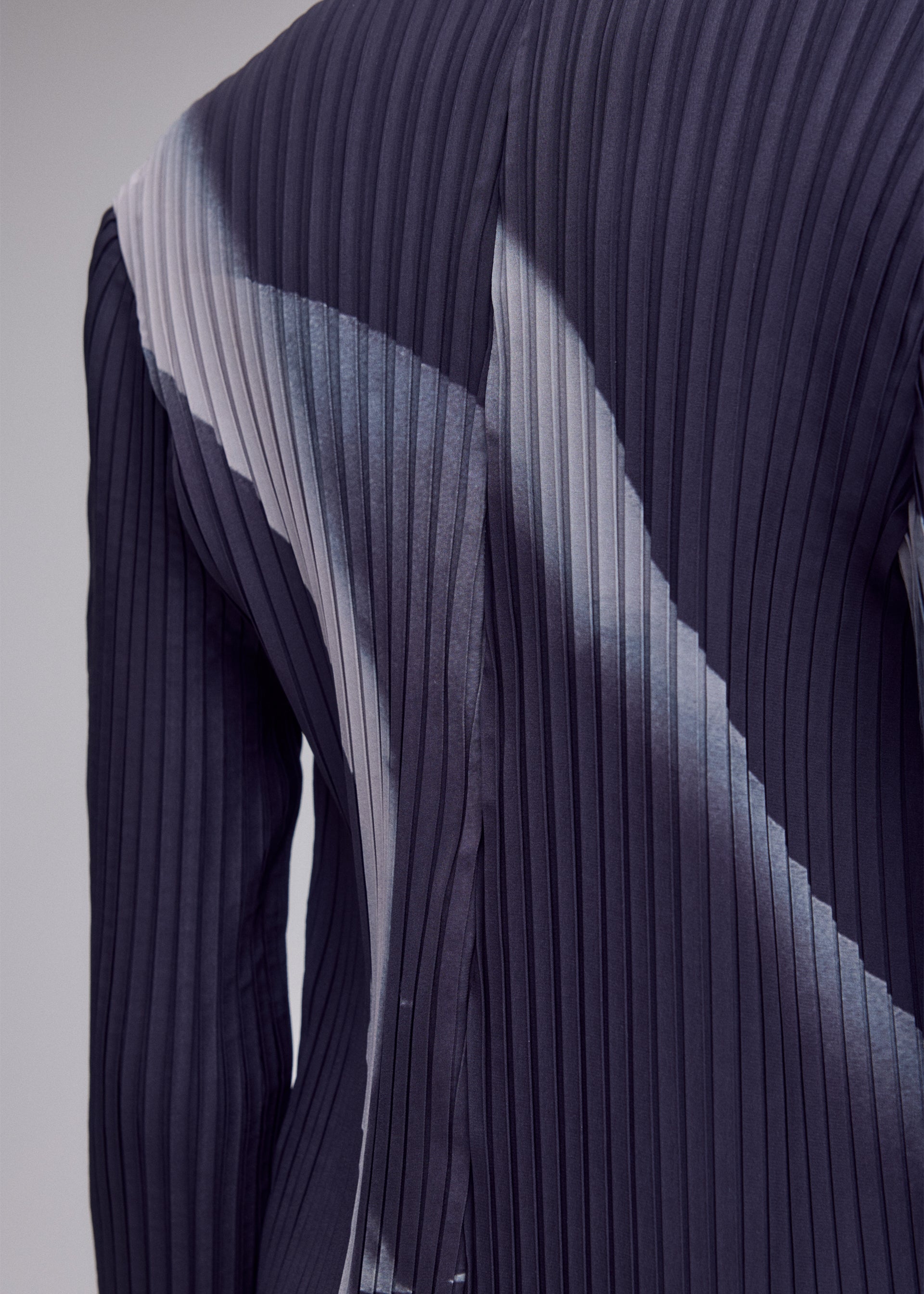 Zoomed view A close-up of the HANDS OF LOVER PLEATS fabric by ISSEY MIYAKE, featuring dark polyester crepe de chine with white diagonal patterns. The pleated textile, likely from a fitted piece and photographed from the back or side against a plain background, showcases an original design created through advanced inkjet printing techniques.