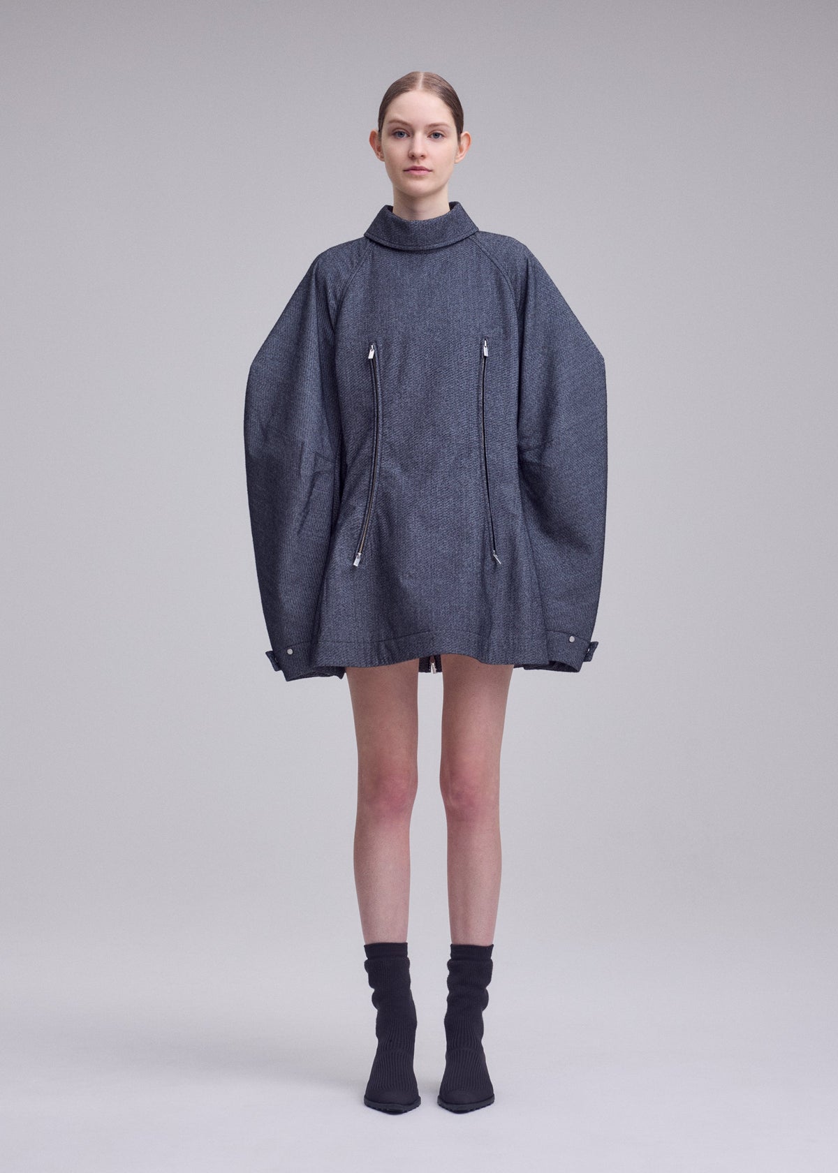 A woman stands in a studio wearing the ISSEY MIYAKE REFLECTION oversized gray jacket with zippers, layered over a silver high-neck tunic and paired with black socks and black shoes. The background is plain and light gray. view 4 of 8