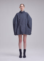 A woman stands in a studio wearing the ISSEY MIYAKE REFLECTION oversized gray jacket with zippers, layered over a silver high-neck tunic and paired with black socks and black shoes. The background is plain and light gray.