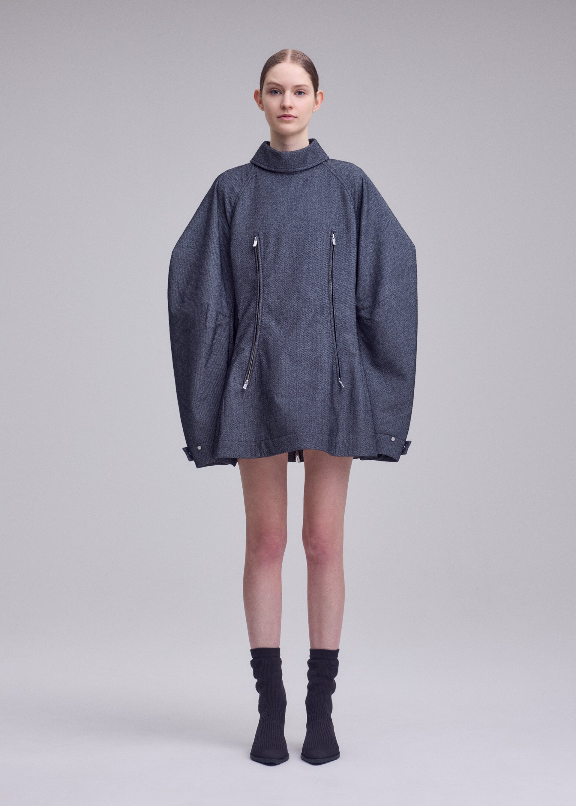 Zoomed view A woman stands in a studio wearing the ISSEY MIYAKE REFLECTION oversized gray jacket with zippers, layered over a silver high-neck tunic and paired with black socks and black shoes. The background is plain and light gray.